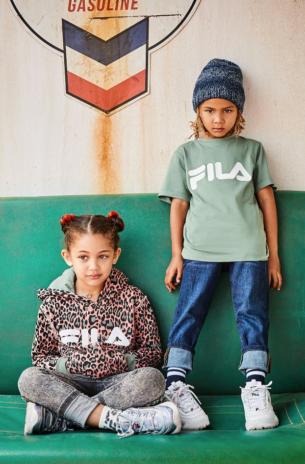 A waist-up photo of a skier dressed in a ski gear standing on a snowy mountain. The background is blurry and white, with a snow-covered tree-lined mountain barely visible. The skier is wearing a bright red jacket with a hood, a brimmed cap, sunglasses, bright blue and yellow gloves, and dark overalls beneath the jacket.